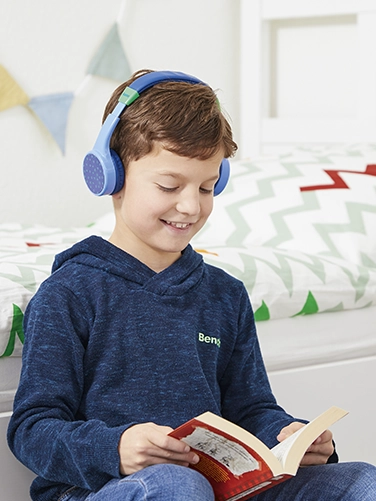 Ein Junge sitzt in seinem Zimmer und liest ein Buch, während er den Hama Bluetooth®-Kinderkopfhörer "Teens Guard" trägt