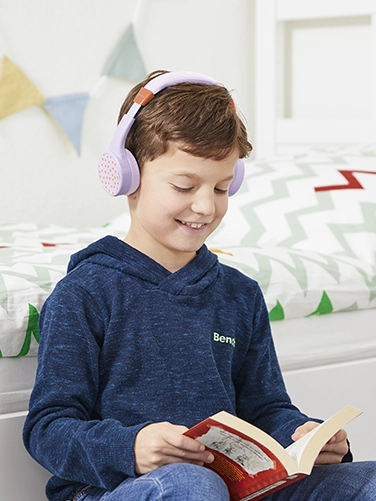 Ein Junge sitzt in seinem Zimmer und liest ein Buch, während er den Hama Bluetooth®-Kinderkopfhörer "Teens Guard II" trägt
