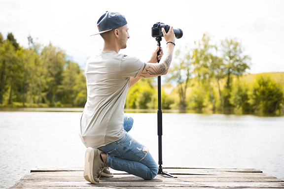 Ein junger Mann kniet auf einem Steg am See und fotografiert die Szene mit seiner Spiegelreflexkamera, die auf dem Hama Monostand "Smooth" befestigt ist