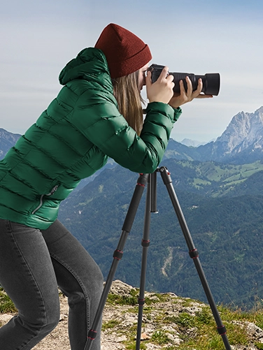 Junge Frau ist mit Jacke und Mütze bekleidet auf einer Anhöhe in den Bergen und fotografiert mit einer Spiegelreflexkamera, die auf dem Hama Dreibeinstativ "Ramble II Duo Carbon" montiert ist